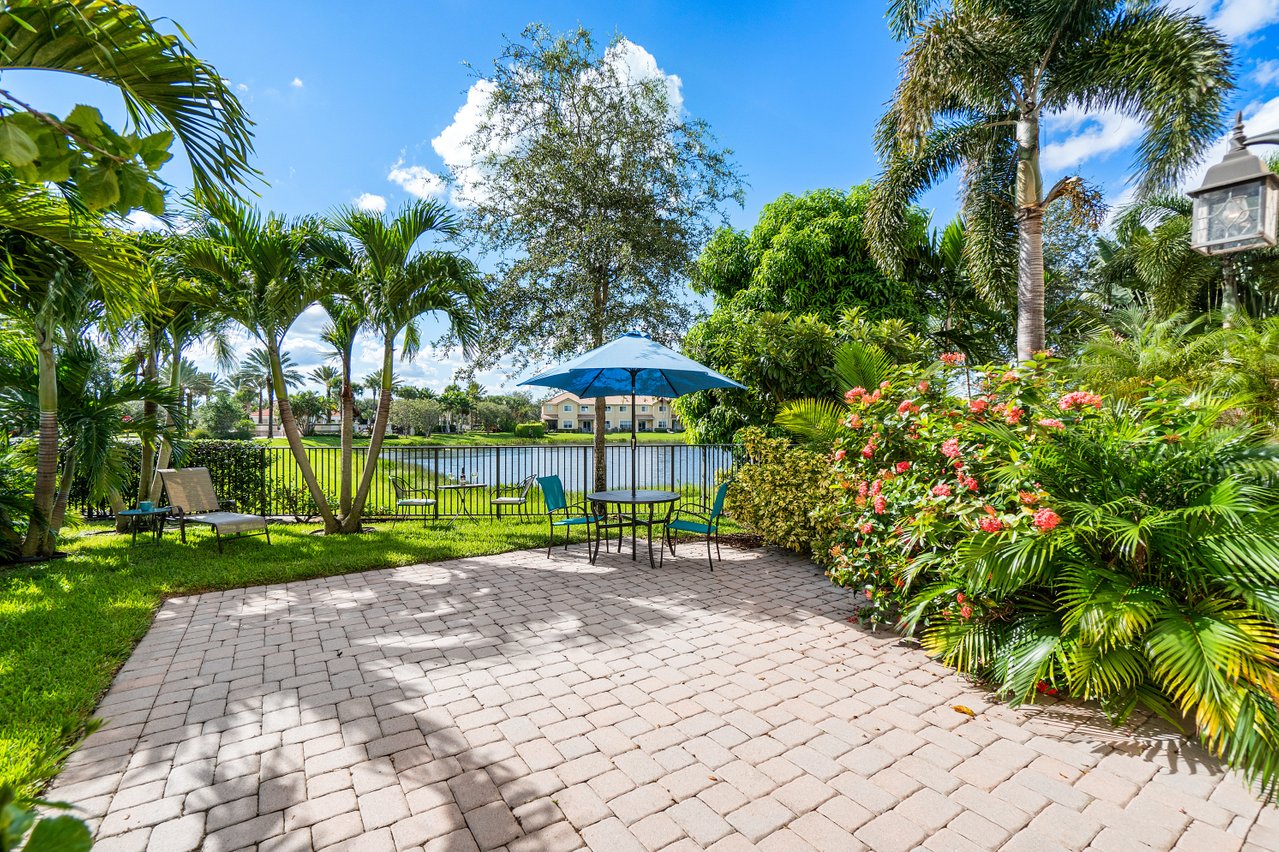 Brick-paver patio with a blue umbrella table overlooking a lake, framed by palms and flowering shrubs, in Palm Beach Gardens, Florida
